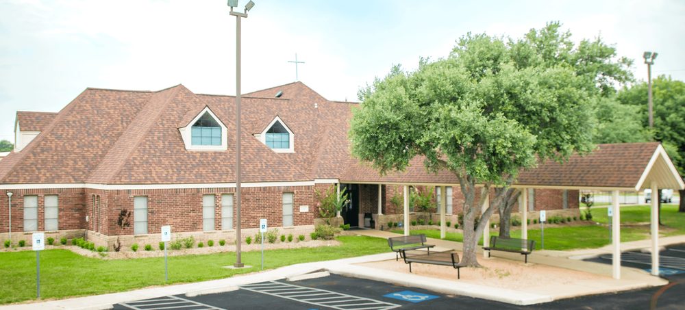 The Westside Baptist Academy campus: a single-storey red brick building with a covered walkway along the front, a large oak tree, and visitor parking directly in front of the entrance.